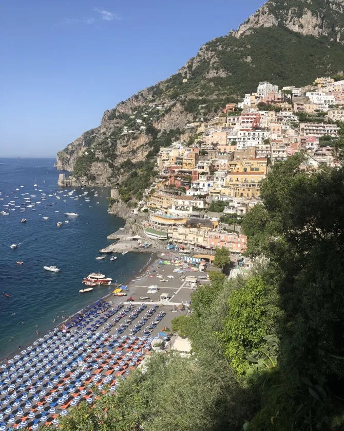 A beautiful view of an Italian beach with a uniform array of beach umbrellas, boats in the water and a colorful coastal town nestled into the green and rocky mountain. 