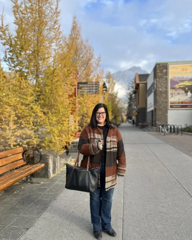 A woman posing with a leather bag in hand on a sidewalk. There are trees and a bench in the background, as well as a building in the far right corner. 