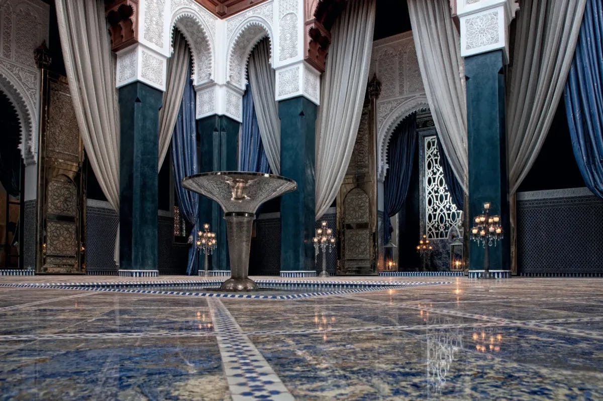 Intricately designed tiles lead up to a center, brass fountain in the middle of an exceptionally ornate Riad-style room. Along the walls, elegantly etched blue and white columns stand, with drapes crossing between.