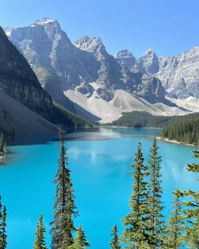 A view of a bright blue lake surrounded by pine trees and large rocky mountains. 