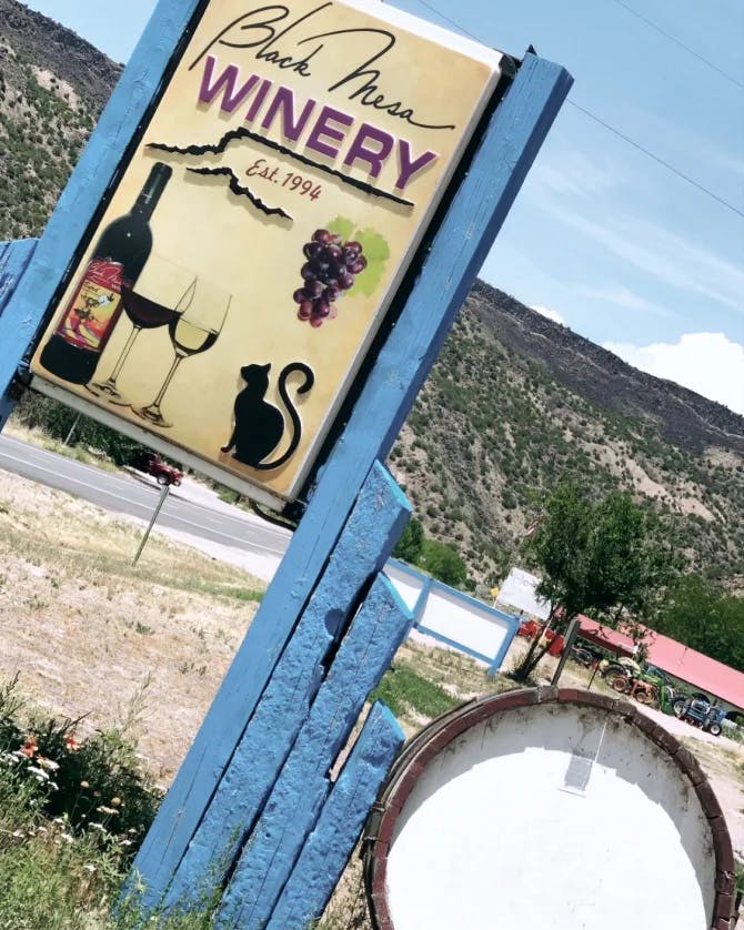 A blue sign for a winery with graphics of a wine bottle, two wine glasses, a black cat and a bundle of grapes. It's placed outside in front of a tree-covered canyon. 