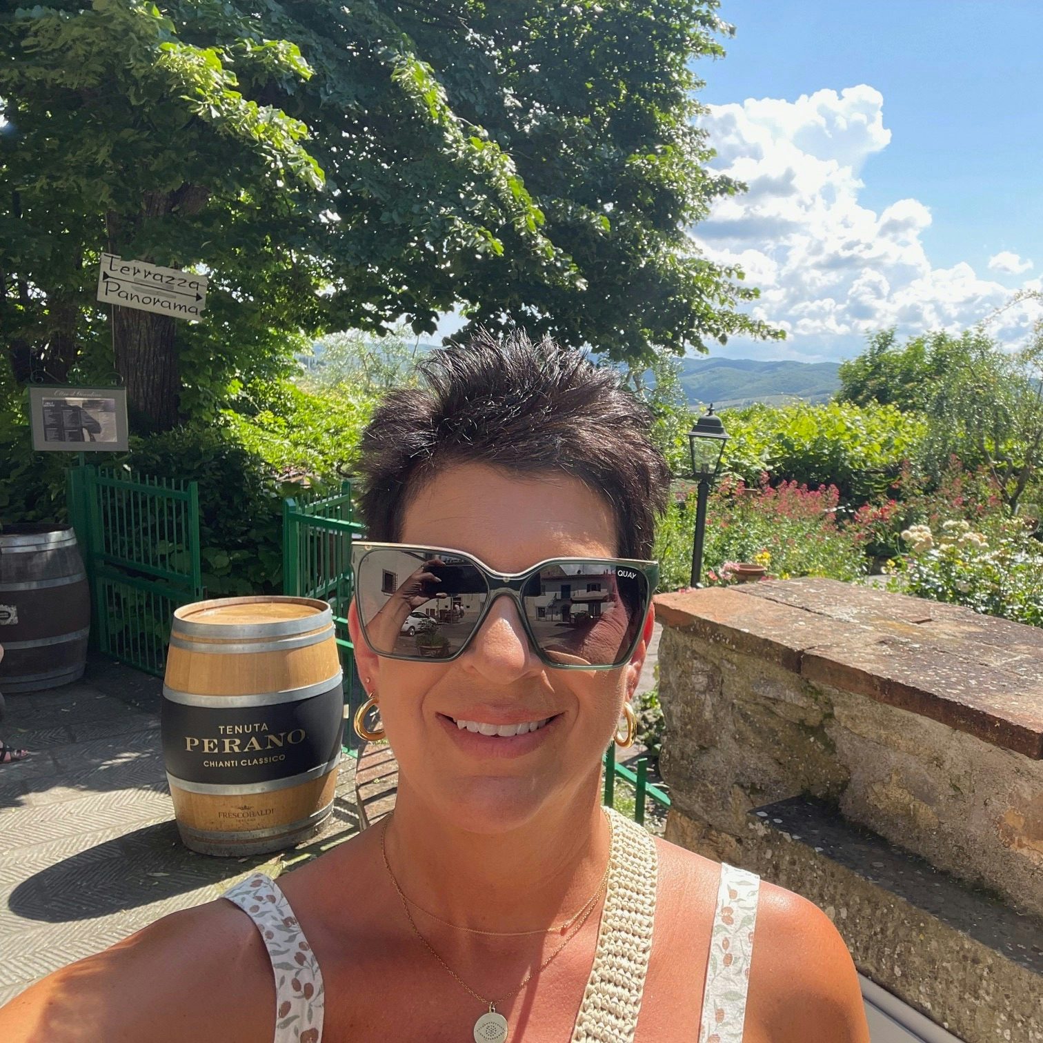 A selfie of Barbara Fodor in a white tank top at a vineyard in front of a barrel that says "Tenuta Perano Chianti Classico," a sign that says "Terrazza Panorama" and green hills. 