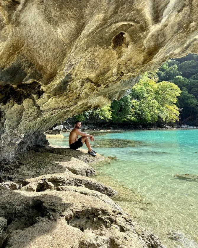  A man sitting inside of a rocky cave near a bright, blue cove full of crystal-clear water. There is a bright green tree in the background too. 