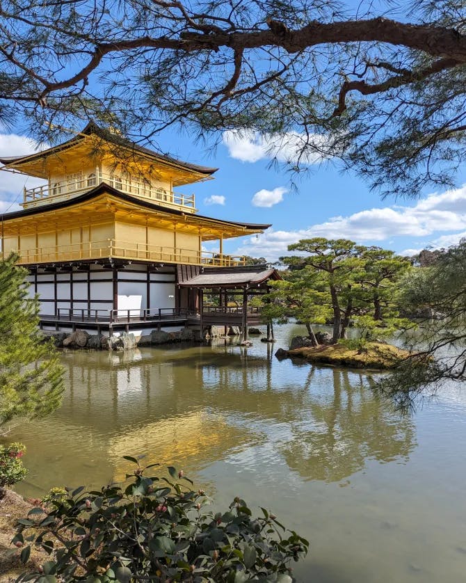A beautiful yellow temple near a lake outside. There are trees and rocks in the surrounding area. 