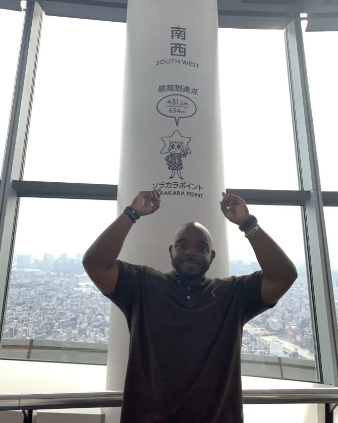 Picture of Harold standing in front of a pole with Japanese writing on it with the view of a skyline in the background. 
