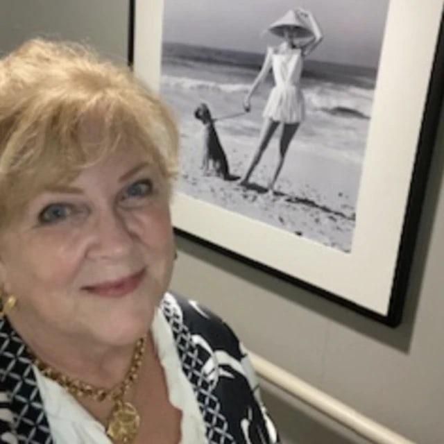 A woman with blonde hair posing for an image with a black and white piece of art in the background showcasing a woman posing on a beach with a dog and leash in hand. 