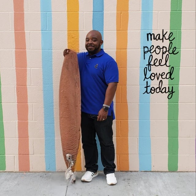 Picture of Harold posing in front of a wall painted in colorful stripes with the quote "Make people feel loved today" written on it in cursive. 