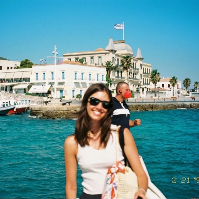 Picture of Alexandra wearing a white top and smiling in front of a blue body of water and white buildings along a coastline. 