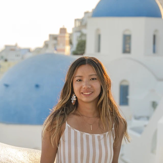 Emma in a white and pink tank top standing in front of blue and white buildings on the coast of Santorini.