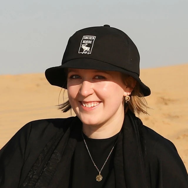 An image of Hannah wearing a black t-shirt and black bucket hat smiling for a photo outside in front of sand dunes. 