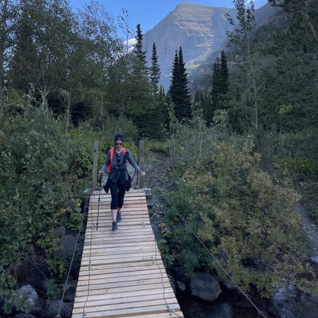 Travel Advisor Kathy Potter hiking across a bridge with mountains and green shrubs around.