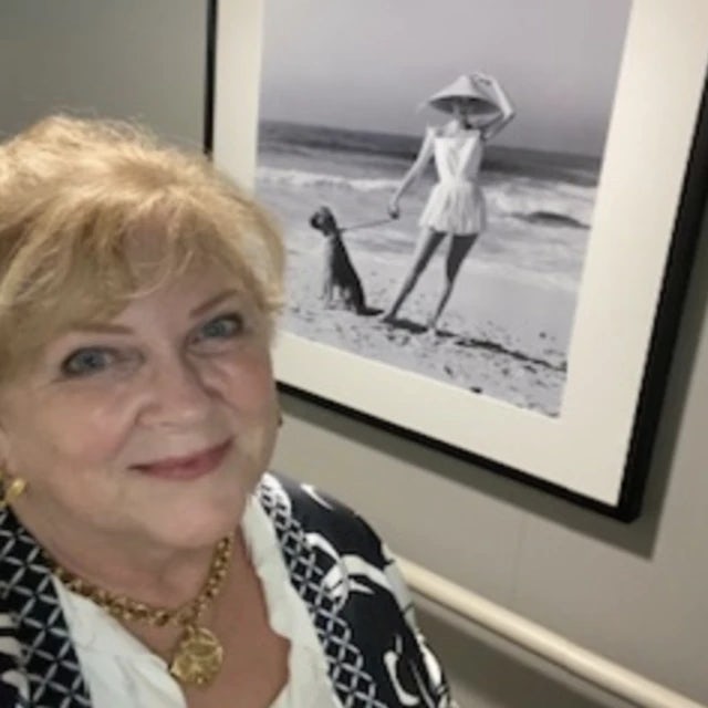 A woman with blonde hair posing for an image with a black and white piece of art in the background showcasing a woman posing on a beach with a dog and leash in hand. 