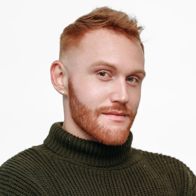 A man with red-colored hair posing in front of a white backdrop with a knit forest-green turtle neck on and his arms crossed in front of his chest. 