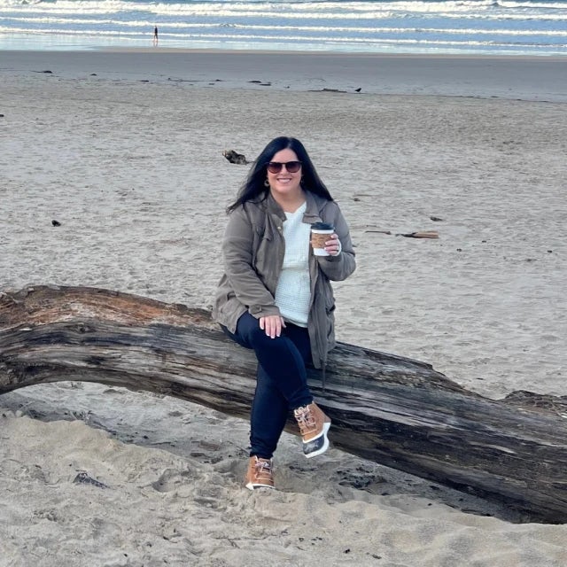 Alicia posing while seated on top of a log on the beach. She is wearing jeans, a white top and a jacket while holding a coffee cup. 